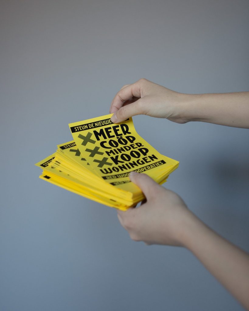Hands holding a stack of yellow flyers reading ‘Meer coöp, minder koop’